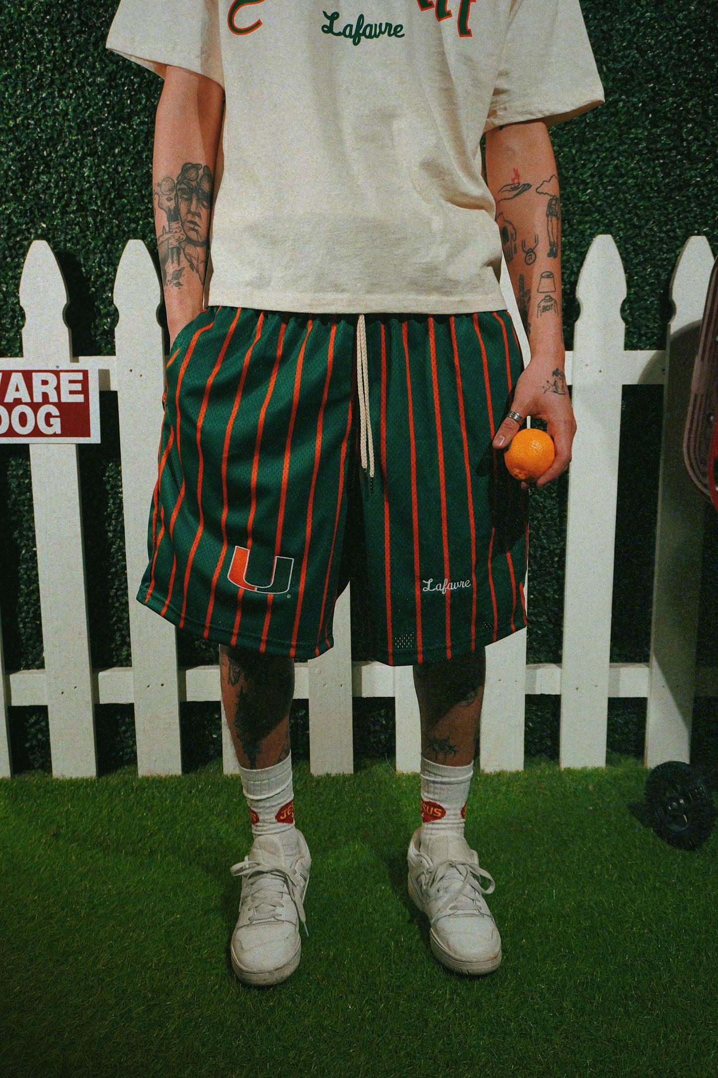 Person wearing a white t-shirt and green striped shorts with a white picket fence and 'Lafavre' sign in the background.