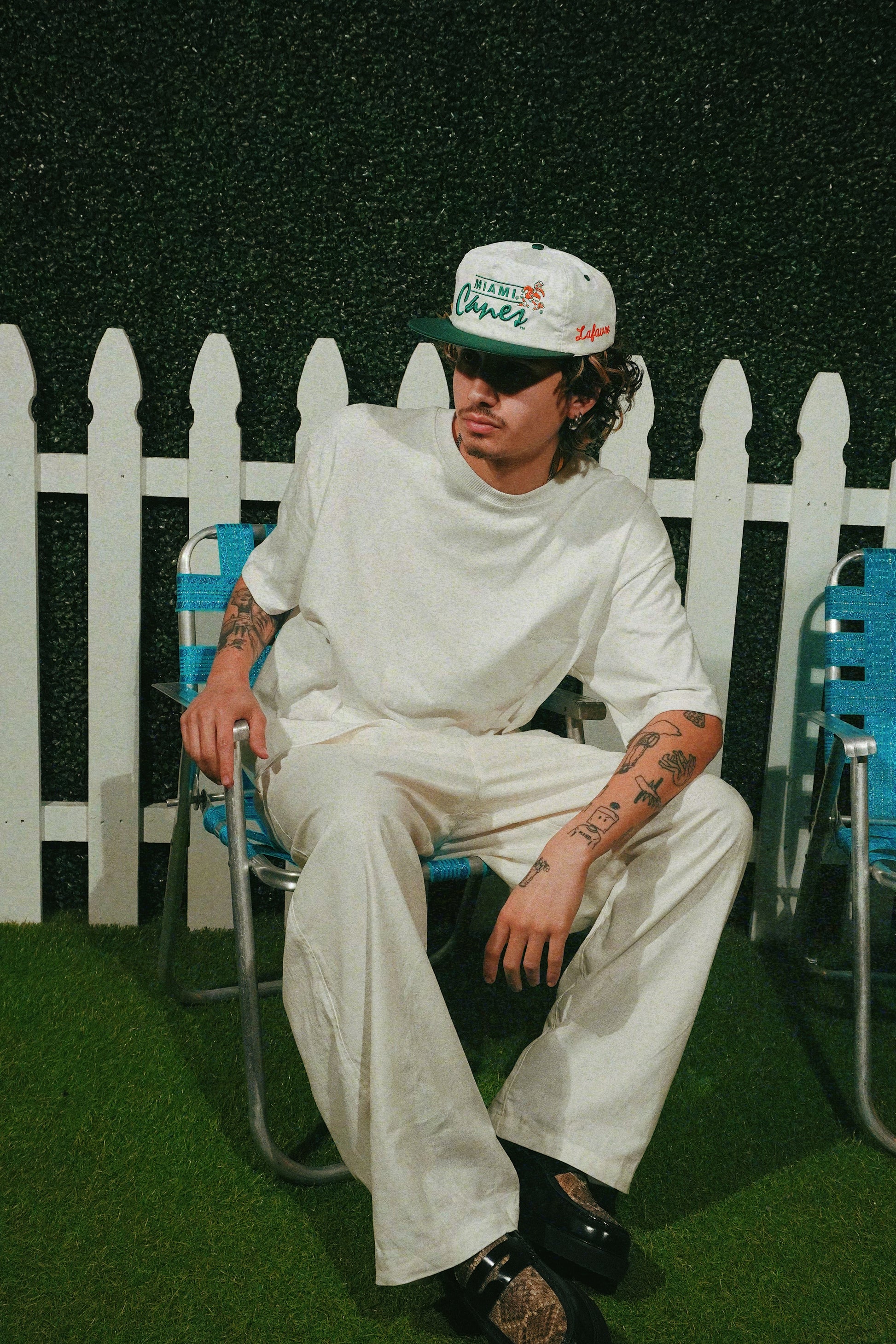 Person wearing a white outfit and cap sitting on a chair in front of a white picket fence.