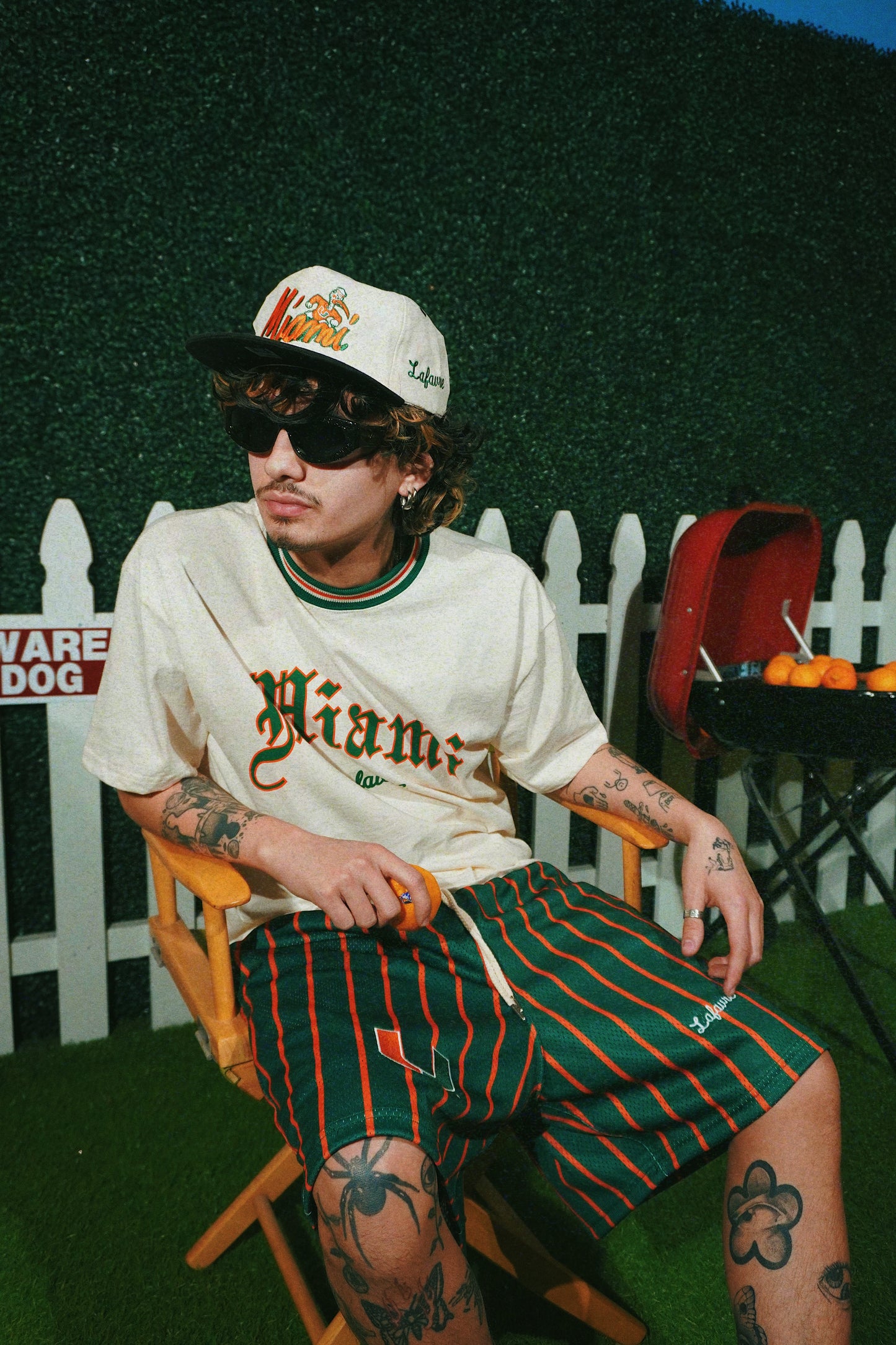 Person wearing a white cap, sunglasses, and a white t-shirt with green and orange accents, sitting on a chair.