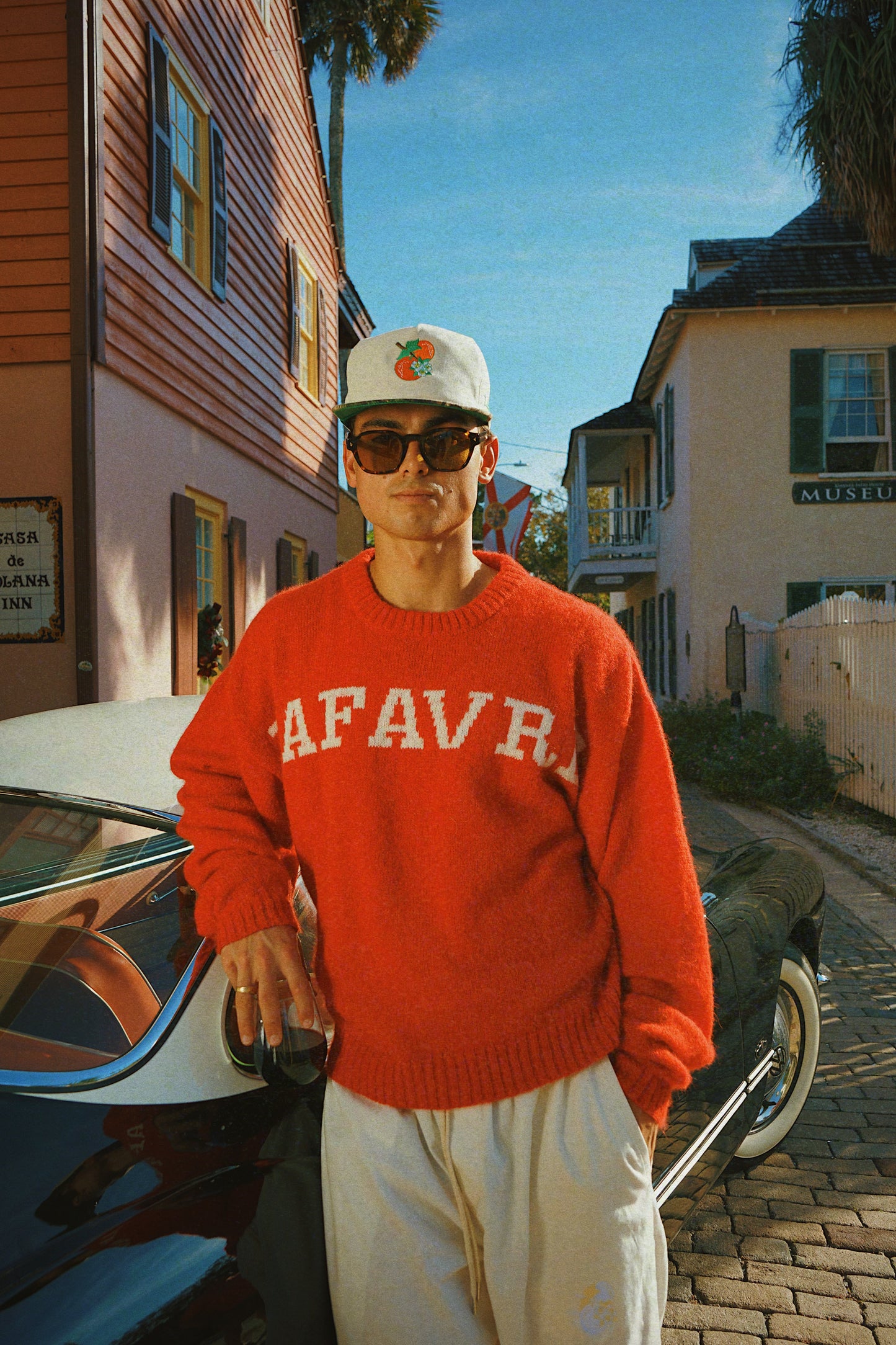 Man wearing a red sweater with 'LAFAVRE' text, white cap, and sunglasses standing next to a vintage car on a street.