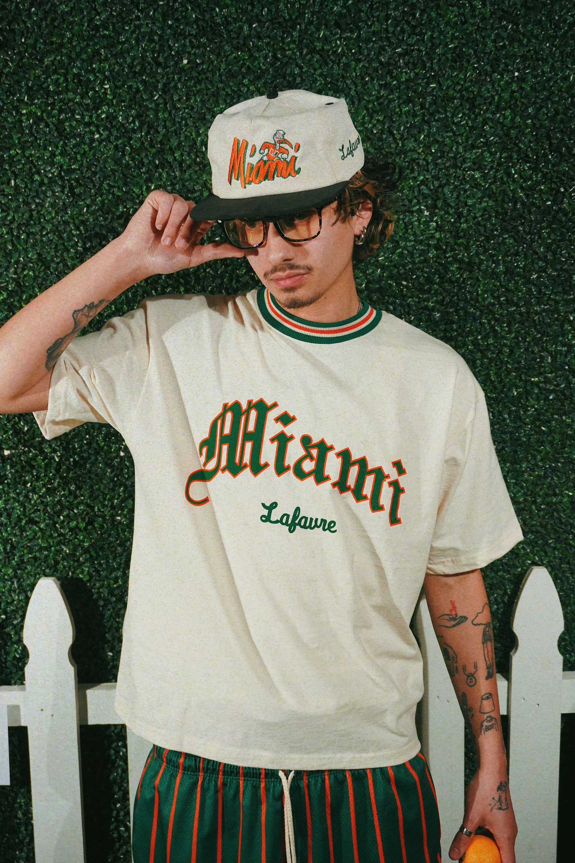 Person wearing a cream-colored t-shirt with 'Miami' print, standing against a green textured wall.