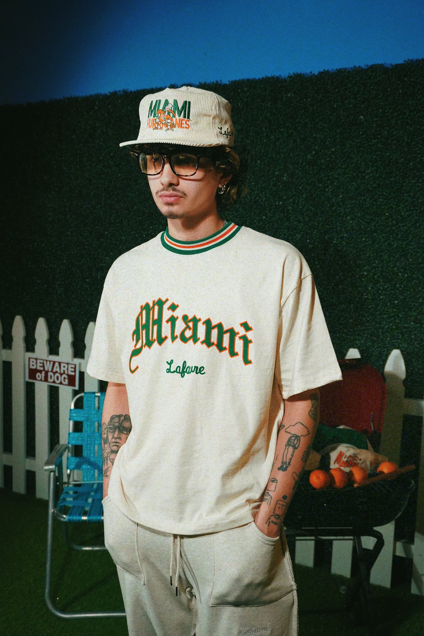 Person wearing a 'Miami' t-shirt and cap in front of a green wall with a white picket fence.