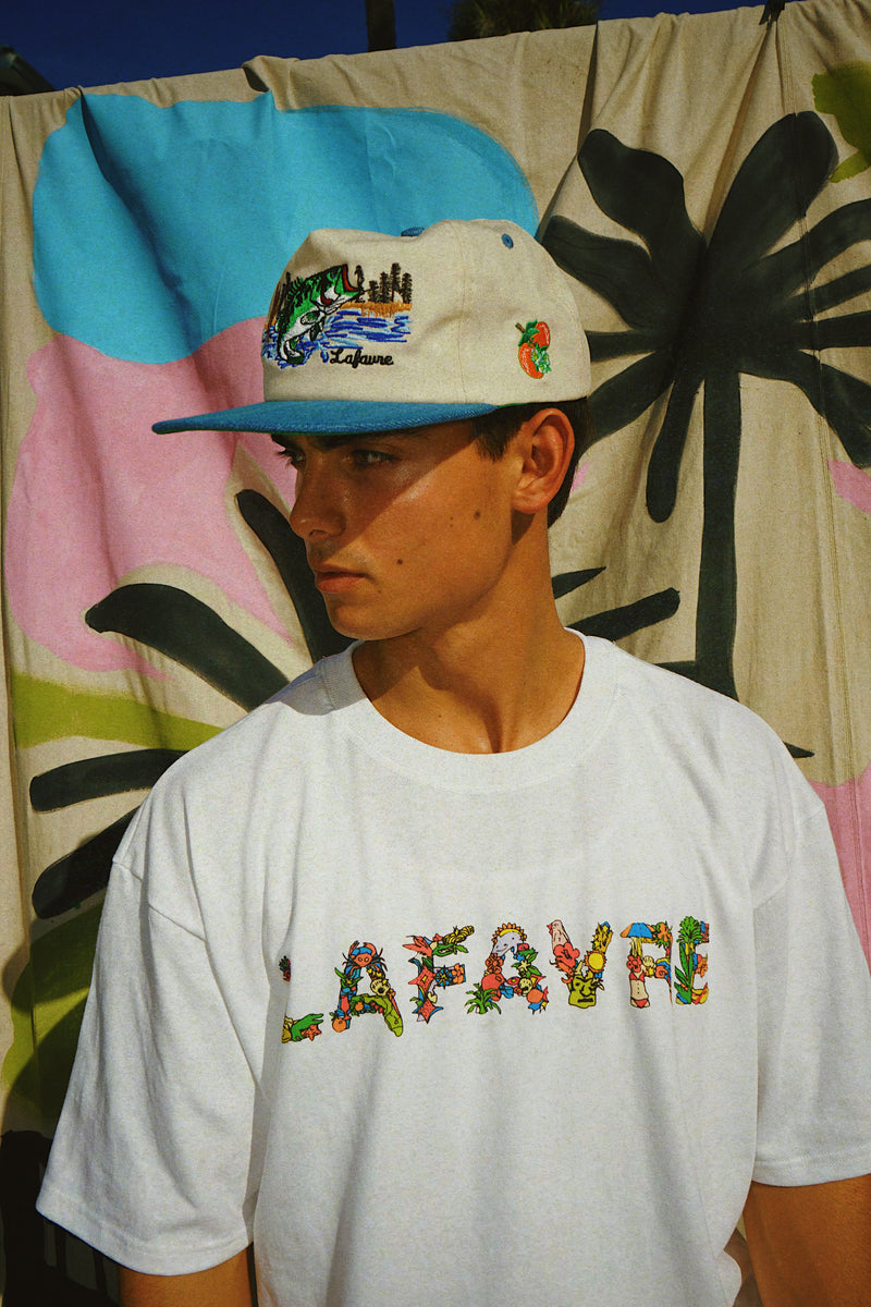 Person wearing a white t-shirt with colorful text and a cap in front of a vibrant, abstract wall.