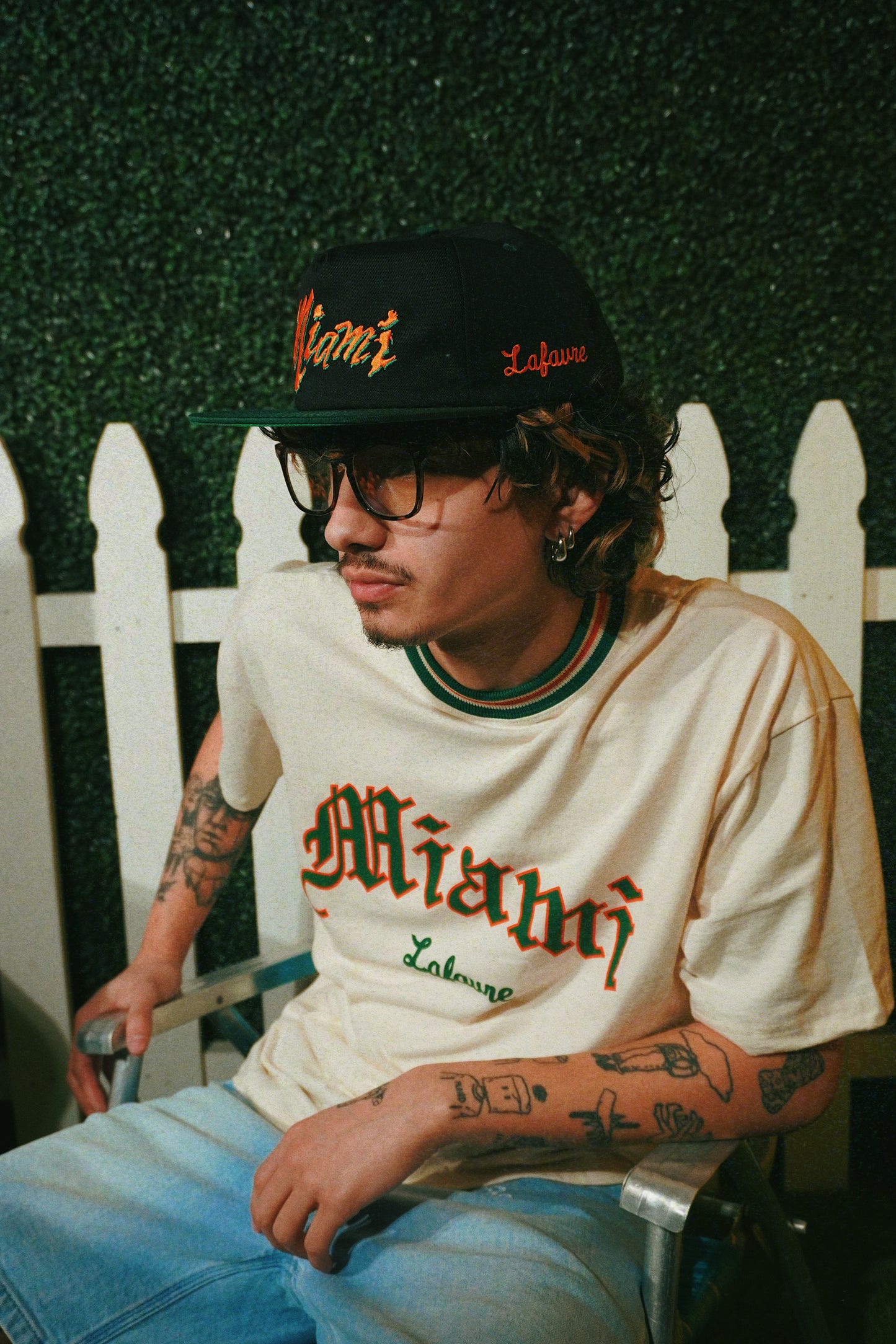 Person wearing a cap and t-shirt with 'Miami' print, sitting on a bench.