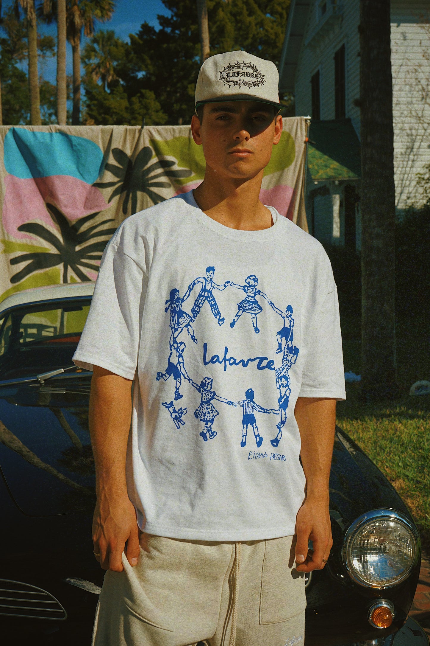 Person wearing a white t-shirt with blue graphics and text, standing in front of a vintage car and colorful towel.