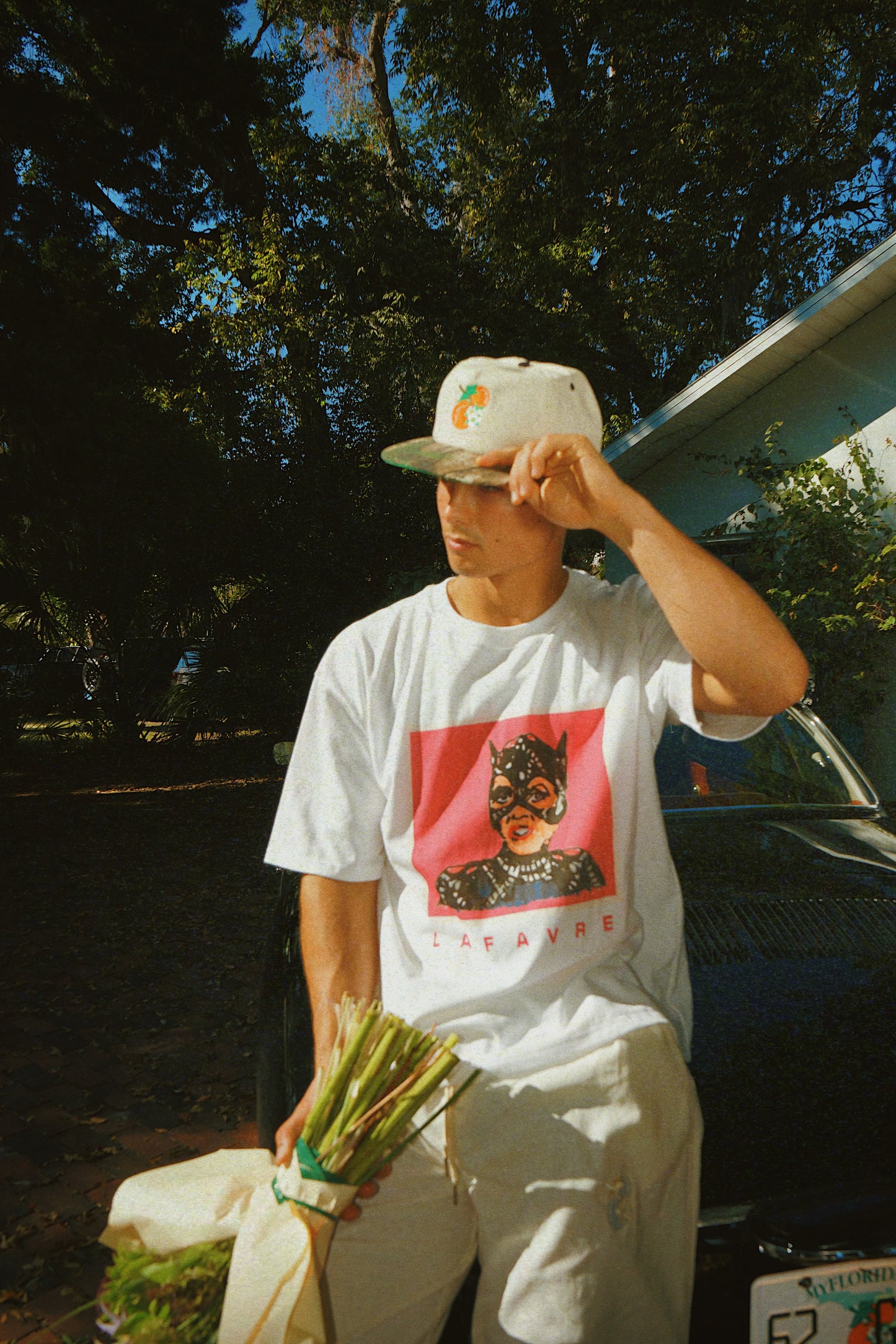 Person wearing a white t-shirt with a graphic design, holding a plant, standing outdoors.