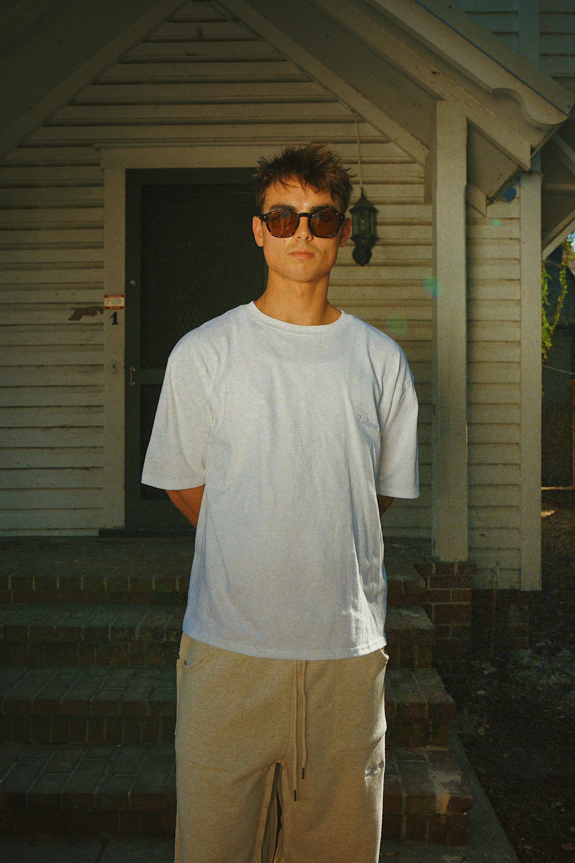 Person wearing sunglasses and a white t-shirt standing on a porch.