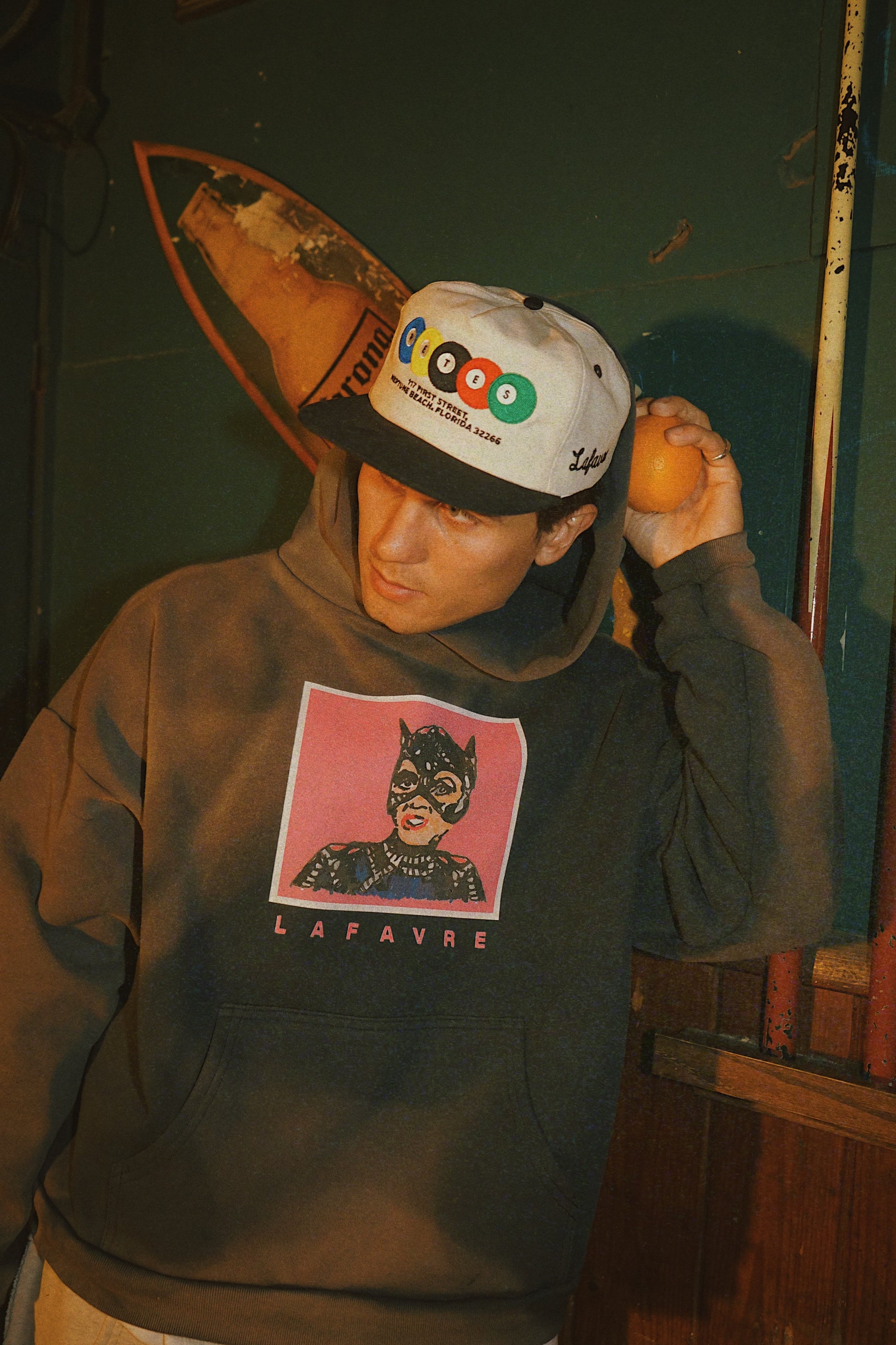 Person wearing a hoodie with a graphic design, holding an orange, against a dark background.