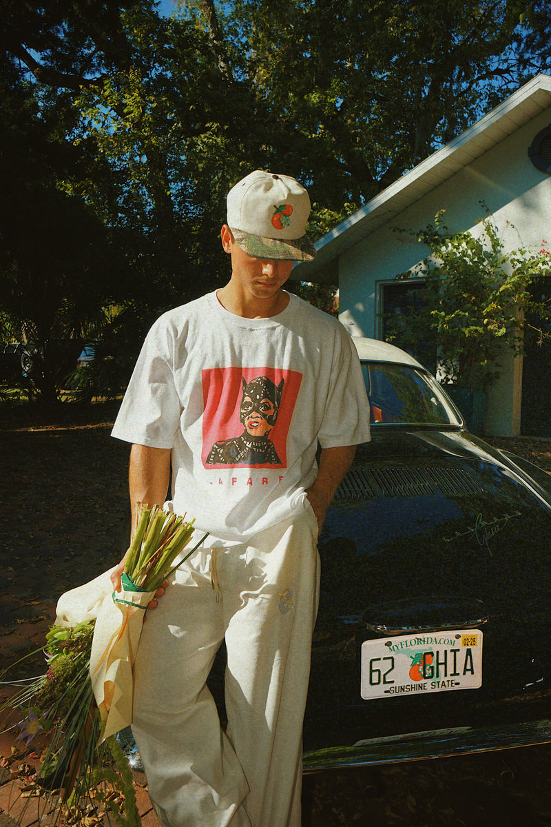 Person standing next to a car holding plants, wearing a white t-shirt with a graphic design and light-colored pants.