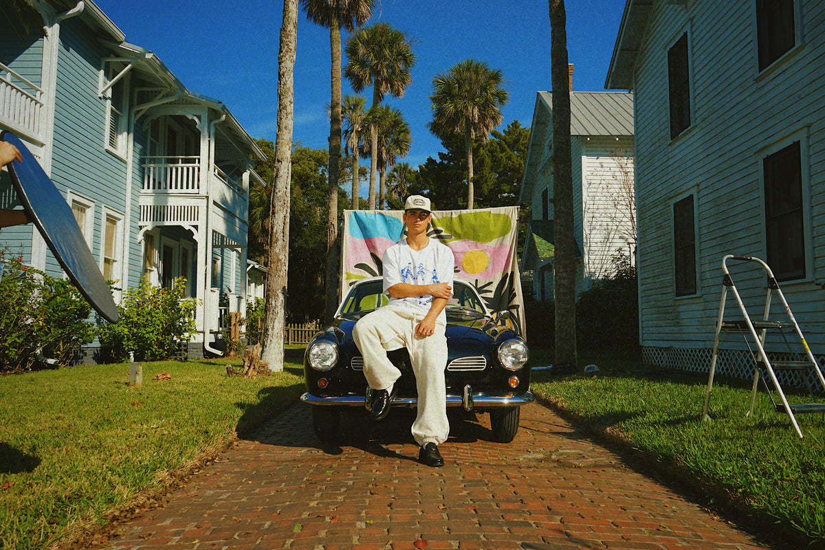 Person sitting on a classic car in front of a house with palm trees in the background