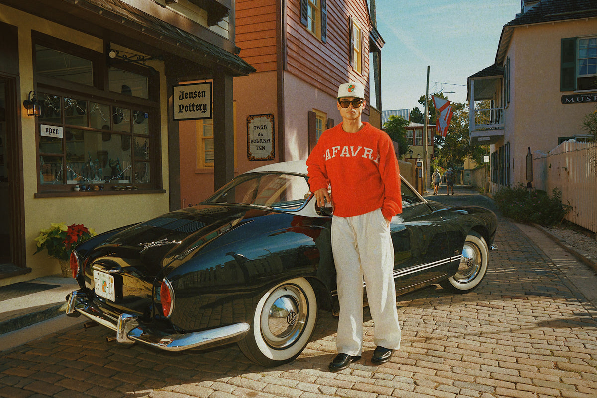 Person in red sweatshirt and white pants standing next to a vintage car on a street.