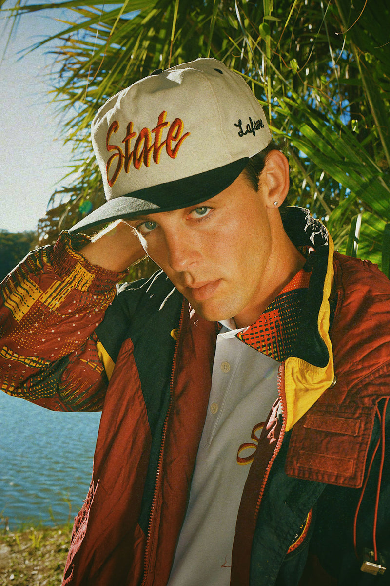 Person wearing a cap with text that says "State" on the front and "Lafavre" on the side, standing outdoors near water and trees