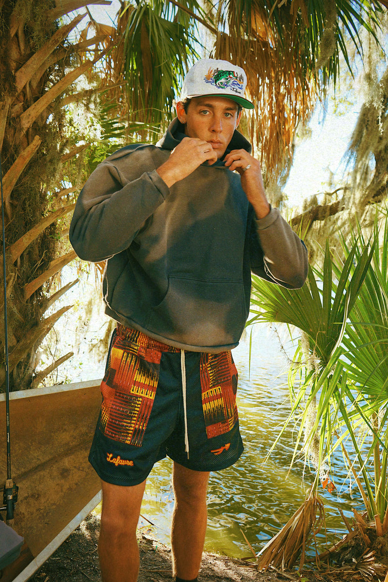 Person wearing a hoodie, Lafavre bass hat, and fire mesh shorts by a body of water with palm trees in the background