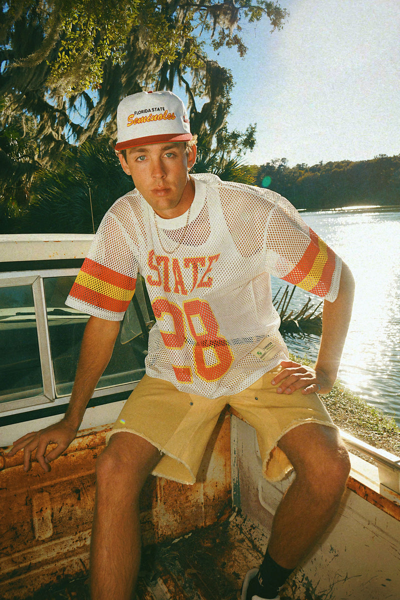Person wearing a white mesh FSU State jersey with orange text and numbers, and a Seminoles cap, sitting on a boat by a body of water.