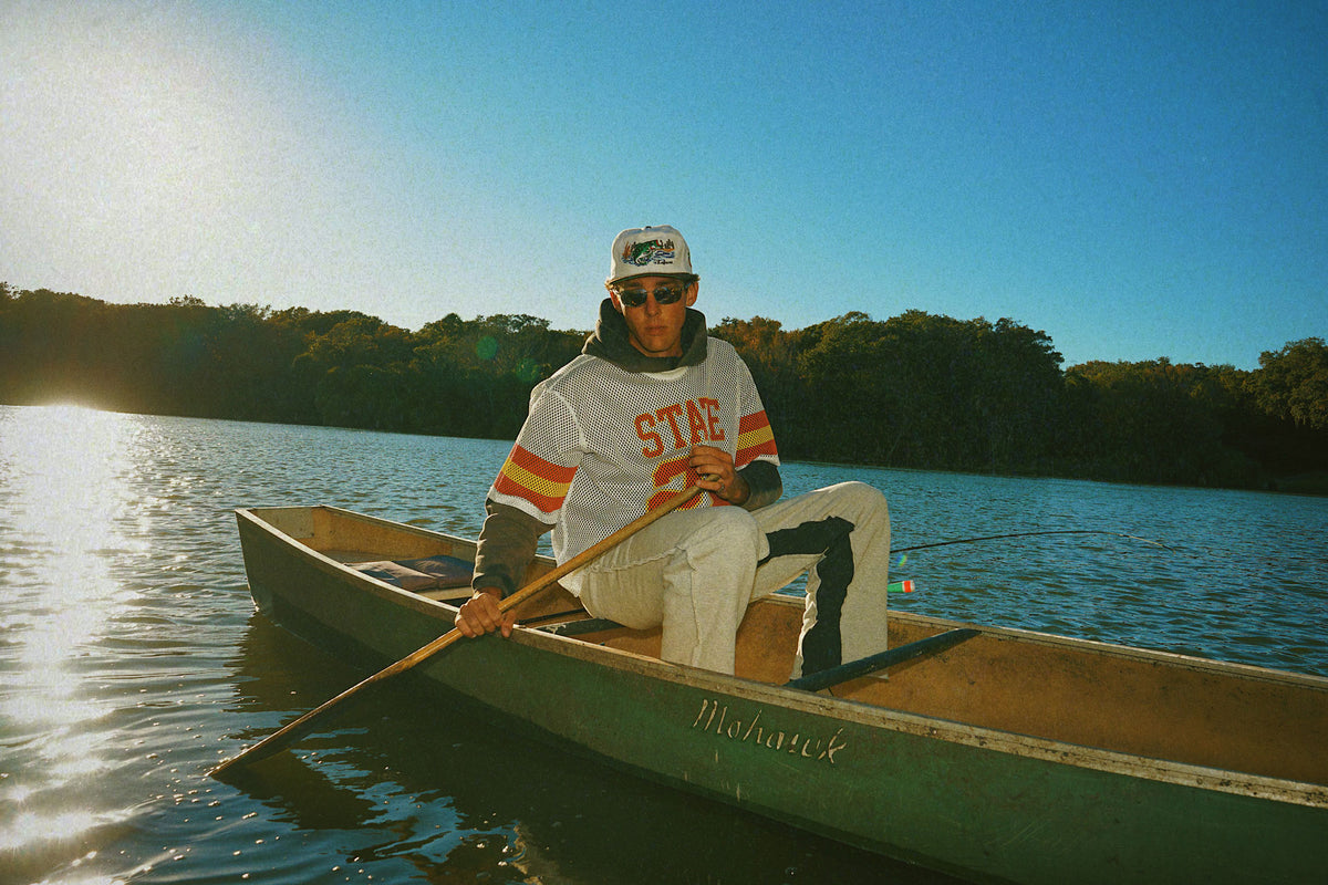Person paddling a canoe on a lake with trees in the background wearing a Lafavre bass hat, and State mesh shirt. 