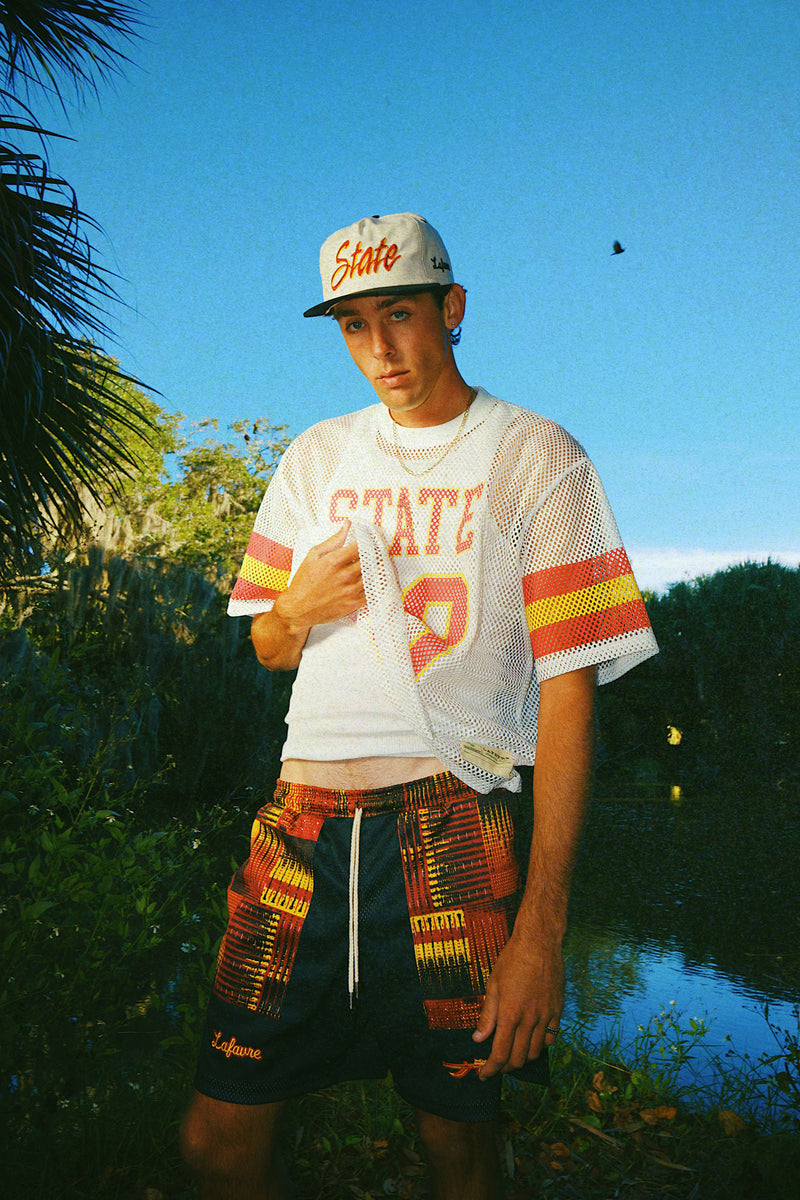 Person wearing a 'State' cap and "State 28" mesh jersey with fire shorts outdoors.