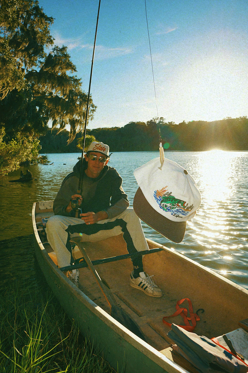 Person sitting in a canoe on a lake with trees and sunlight in the background with a Lafavre bass hat on the end of the rod and reel. 