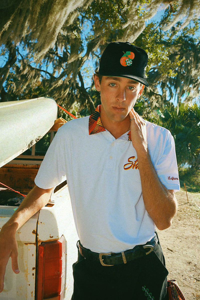 Man wearing a white polo shirt with a State logo and fire pattern collar, black orange blossom cap, and dark pants standing next to a vehicle outdoors. 
